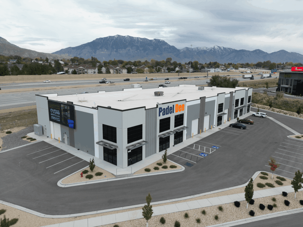 Aerial drone view of Padel Den USA facility in Utah showing modern commercial building with prominent Padel Den signage, blue and orange branding, parking lot with vehicles, highway access, residential neighborhood, and snow-capped mountain backdrop