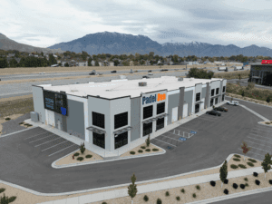 Aerial drone view of Padel Den USA facility in Utah showing modern commercial building with prominent Padel Den signage, blue and orange branding, parking lot with vehicles, highway access, residential neighborhood, and snow-capped mountain backdrop