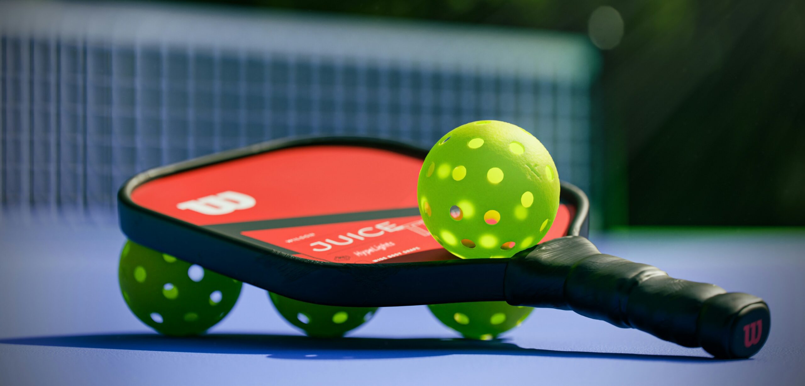 Pickleball equipment close-up: Wilson JUICE paddle with red face and green perforated balls, showing distinctive pickleball gear for padel player comparison