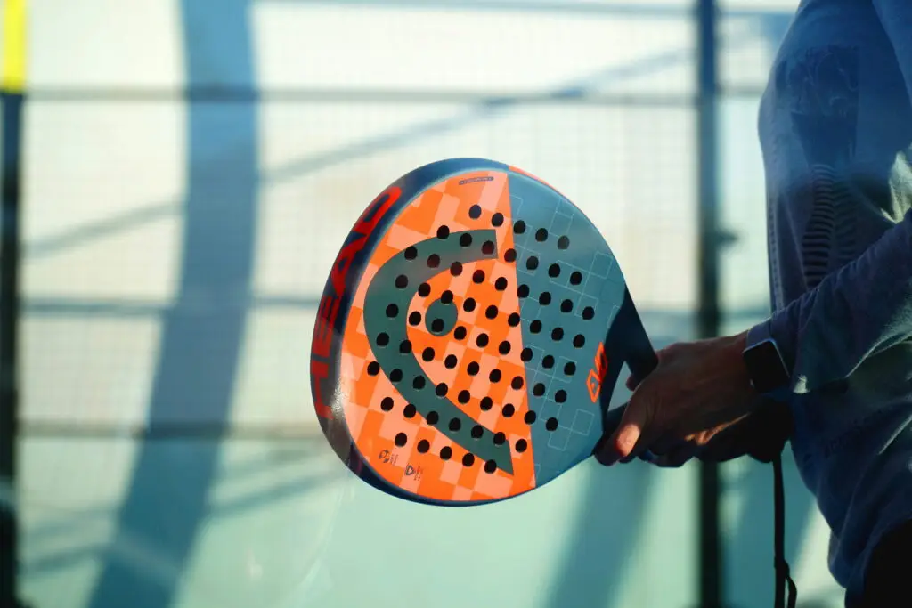 Padel tennis racket close-up held by player at outdoor court in Utah, featuring HEAD branded orange and blue paddle with geometric pattern, demonstrating padel equipment for America's fastest-growing racquet sport