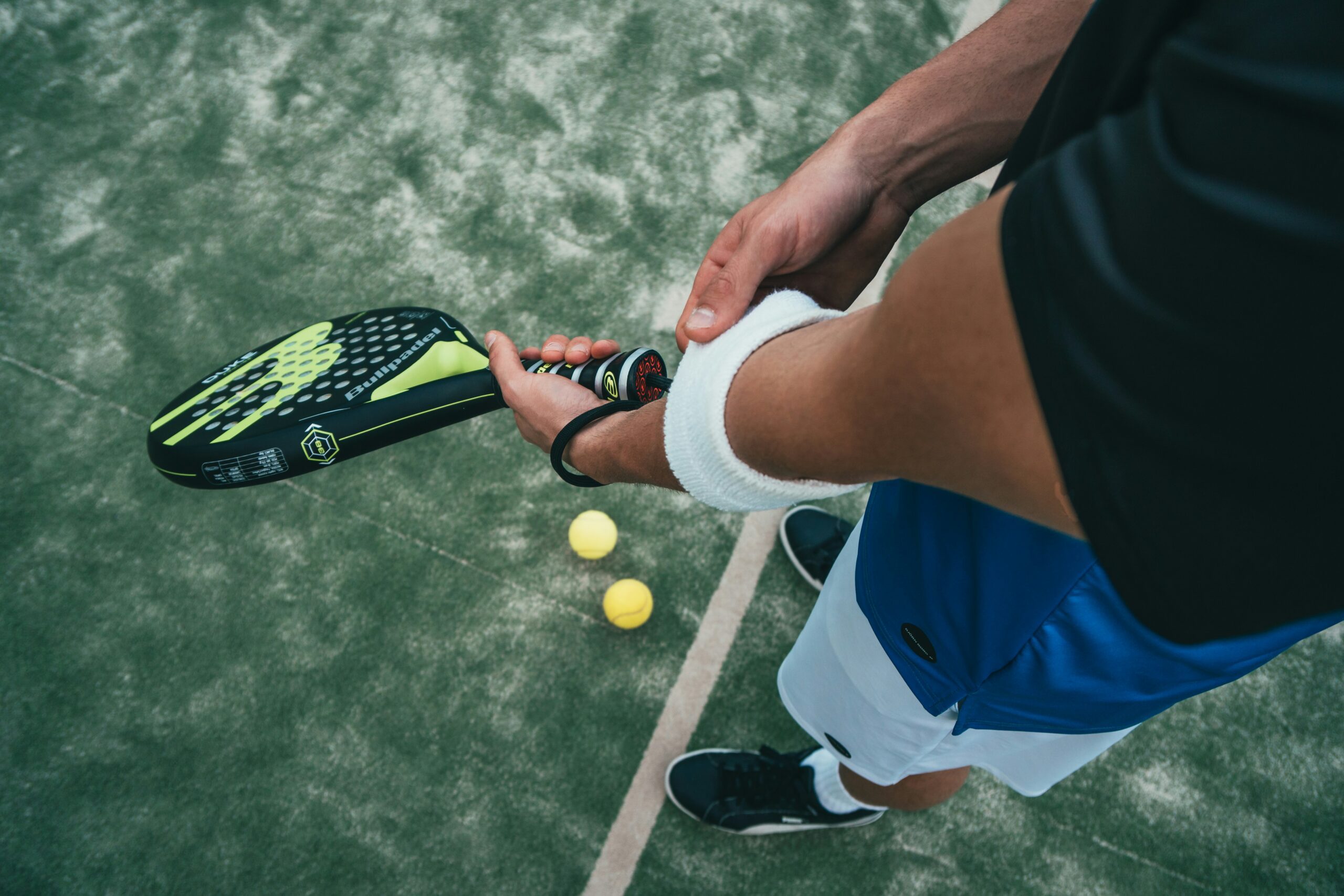 holding racket with both hands in ready position on court, featuring black and neon green paddle and two yellow padel balls on artificial turf, showing proper grip and preparation stance for beginners learning the sport