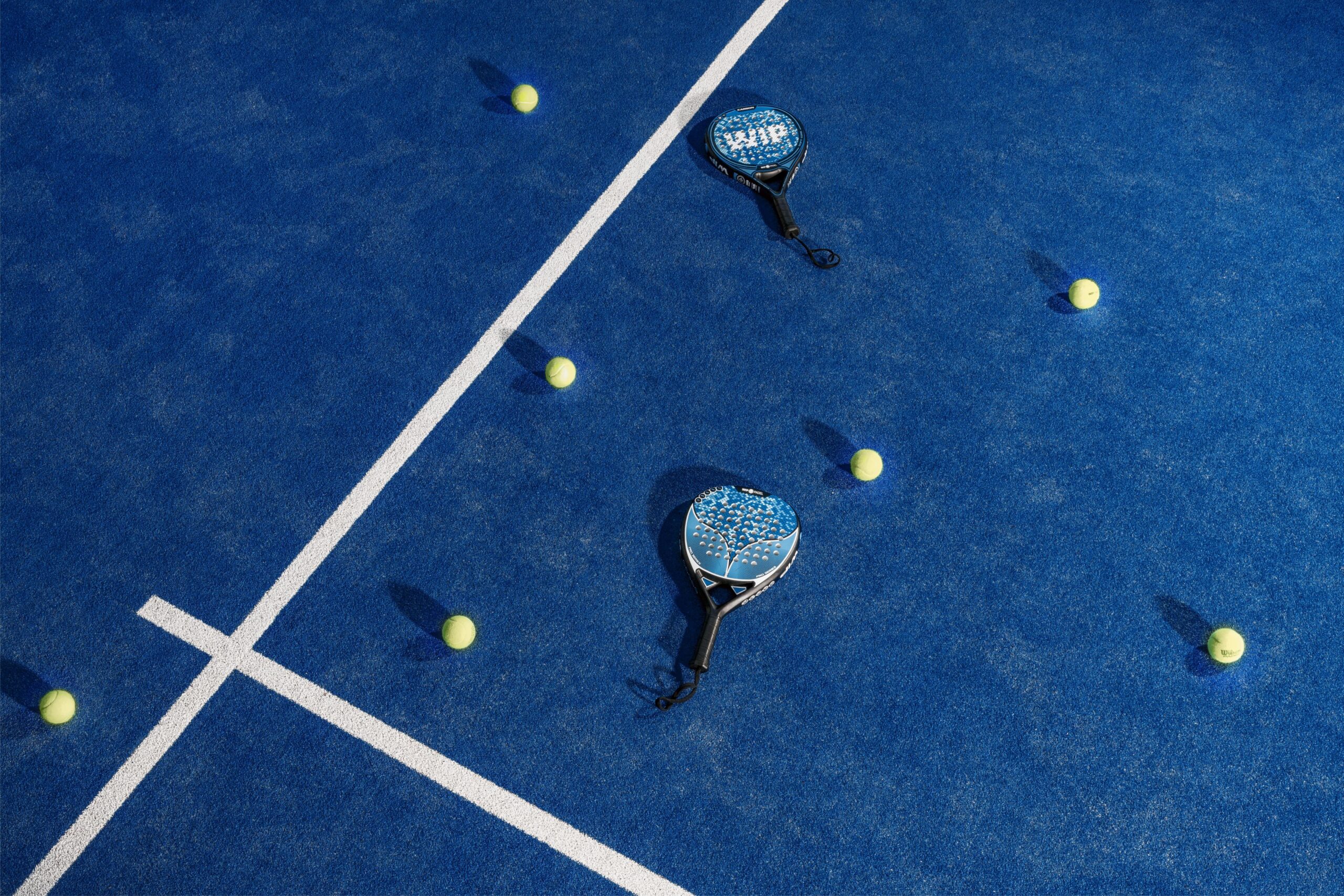 Padel equipment on blue court: Wilson rackets with blue white faces and yellow tennis balls scattered on padel court surface, showing equipment comparison for pickleball players