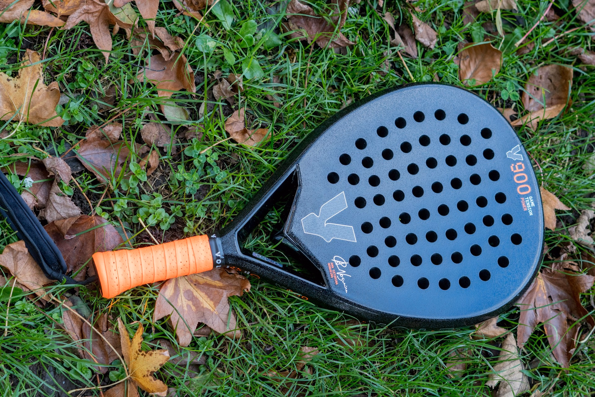 Close-up view of a black V900 padel racket with a neon orange grip resting on green grass and fallen autumn leaves.