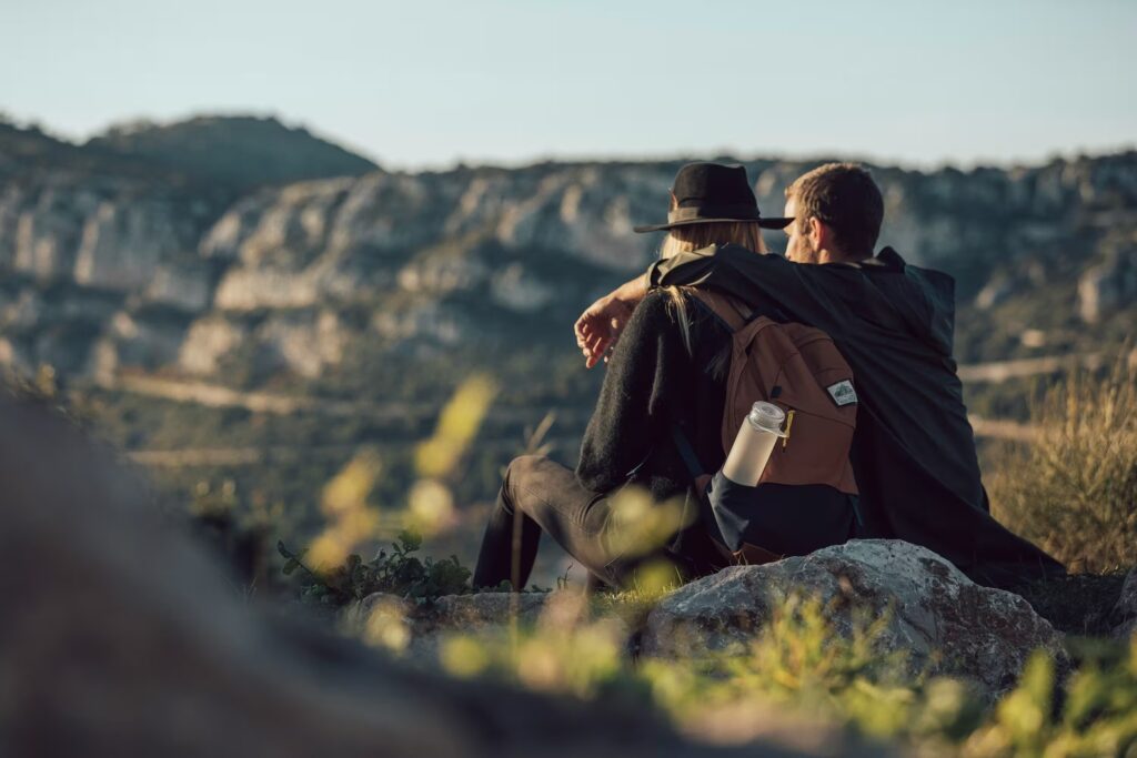 A couple wearing outdoor gear sits close together on a rocky overlook, enjoying a scenic mountain view during an active outdoor date.