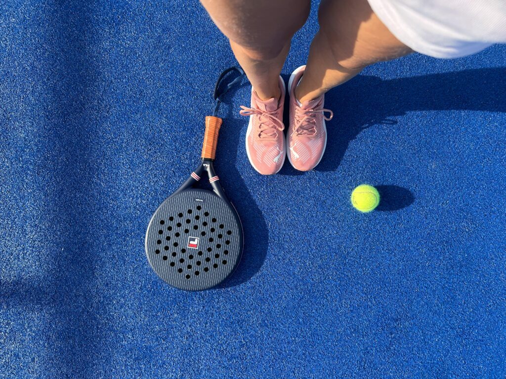 A specialized padel tennis racket and court shoes on a blue artificial turf surface.