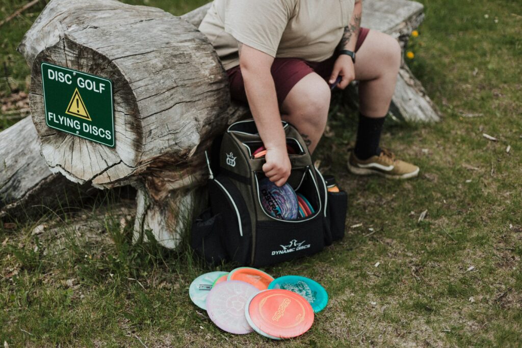 A specialized disc golf backpack sitting on the grass next to colorful flying discs and a rustic log sign reading "Disc Golf".