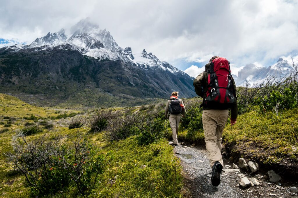 Two hikers with backpacks walking along a scenic trail toward snowy mountain peaks in Utah County, similar to Provo Canyon hikes.
