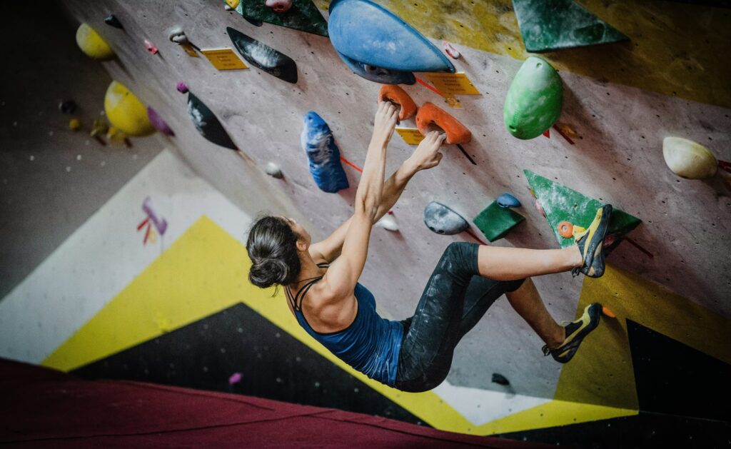 A woman in climbing gear bouldering, scaling an indoor rock wall with bright blue, red, and green holds at The Quarry in Provo.