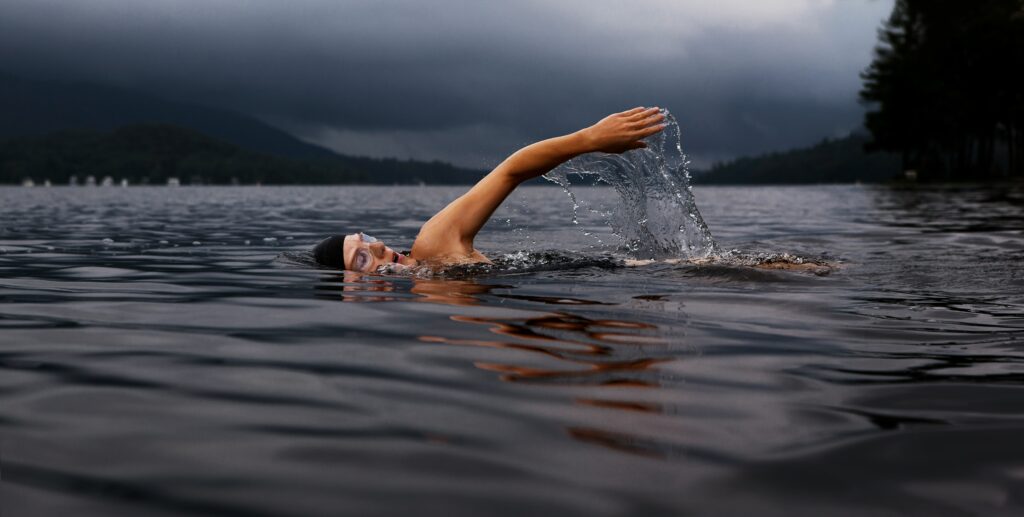 A person swimming in a lake with mountains in the background, showing how swimming is one of the best low impact sports for full body conditioning.