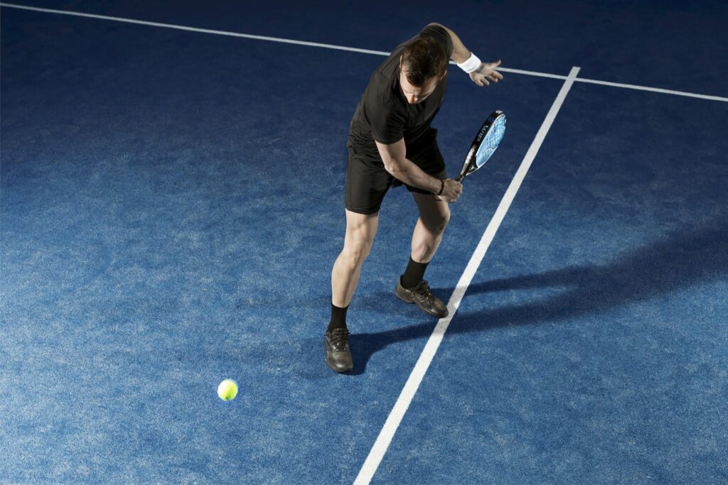 An athletic male player in all-black gear captured mid-swing on a modern blue Padel court, perfectly illustrating how Padel is one of the best low impact sports for sustainable fitness.