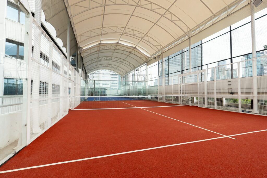An empty, enclosed padel tennis court featuring a red playing surface, glass back walls, and metal fencing.