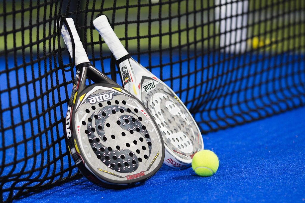Two professional padel rackets and a yellow ball resting against the net on an indoor blue turf court in Utah.