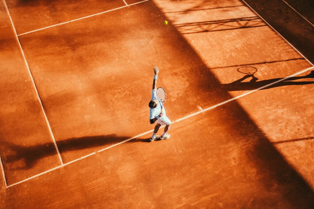 An overhead view of a player hitting a powerful overhand serve on a red clay tennis court, contrasting with the underhand serve used in padel tennis.