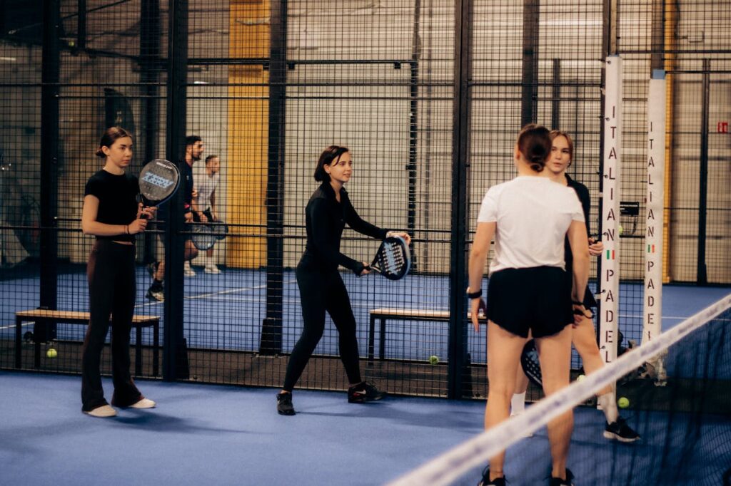 A group of women smiling and communicating at the net during an indoor padel match, highlighting the fun and social atmosphere of the sport.