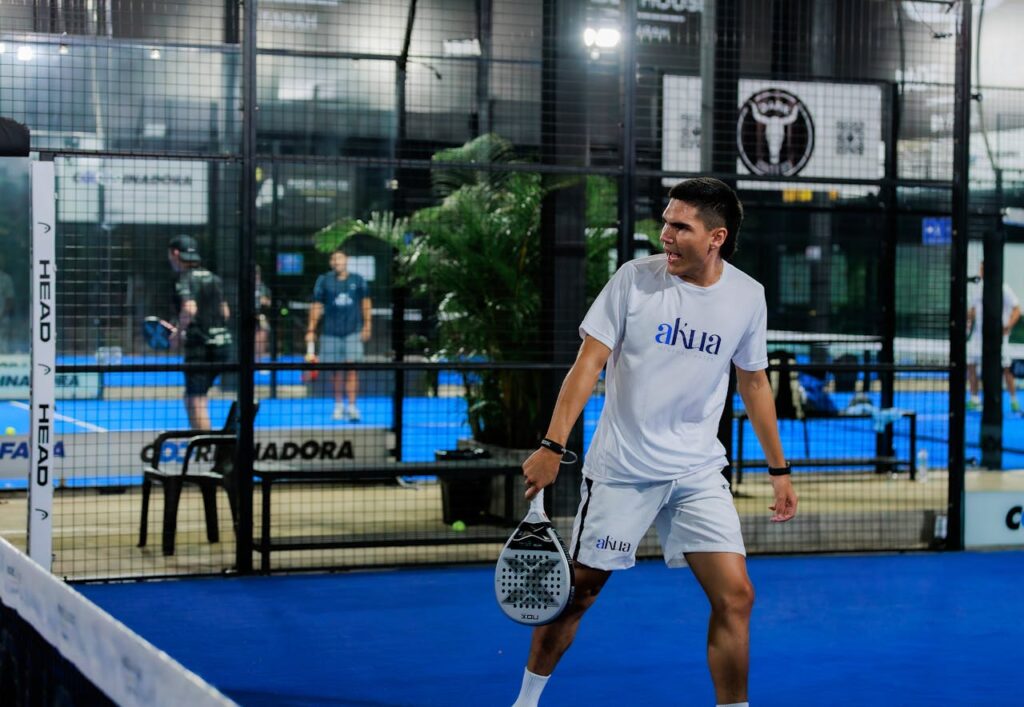 A focused male athlete playing indoor padel, preparing to return the ball after it bounces off the back glass wall.