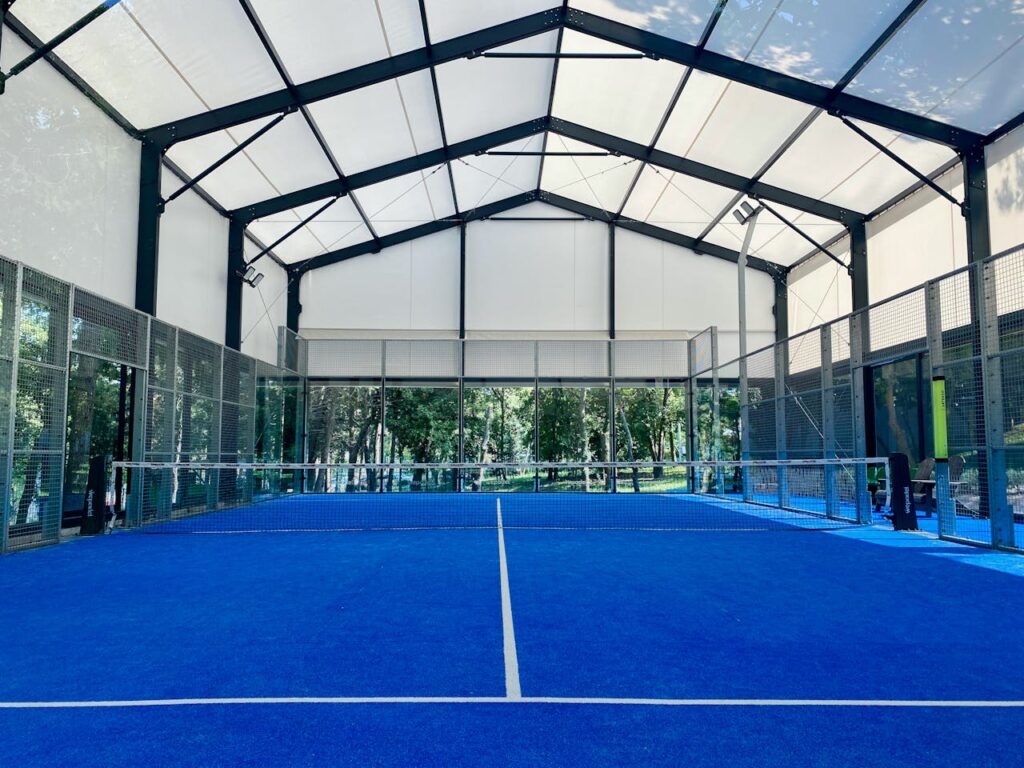 A wide view of a premium indoor padel court with a covered roof and blue artificial turf, offering perfect year-round playing conditions.