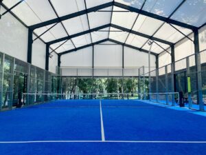 A wide view of a premium indoor padel court with a covered roof and blue artificial turf, offering perfect year-round playing conditions.
