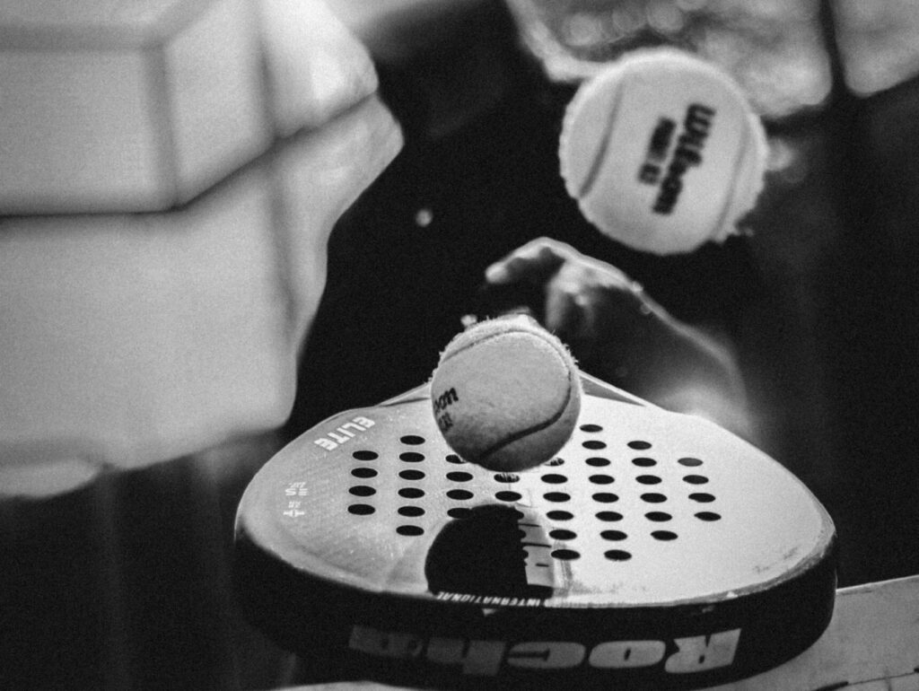 An artistic black and white close-up of a padel racket and ball, showcasing the accessible equipment that makes the sport so popular.