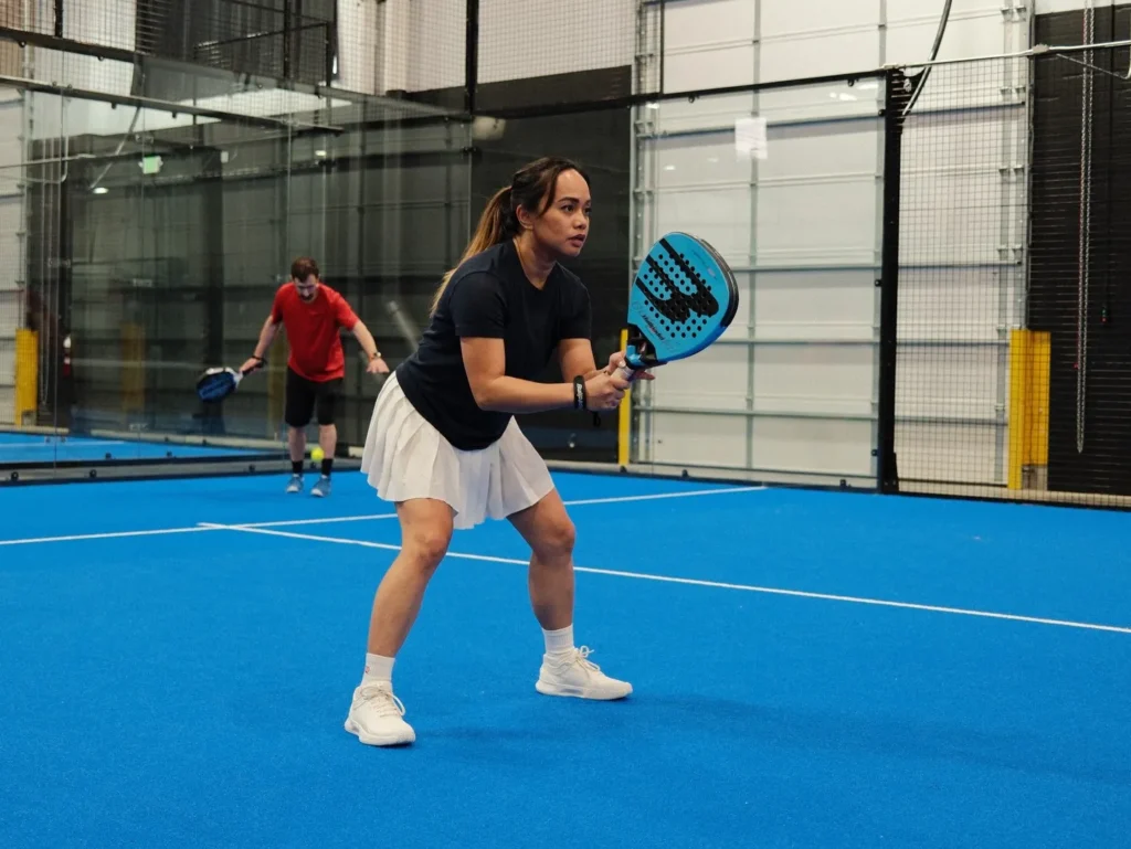 A focused female player waiting to return a serve on the blue turf, showing how fun and accessible it is to play padel in Orem.