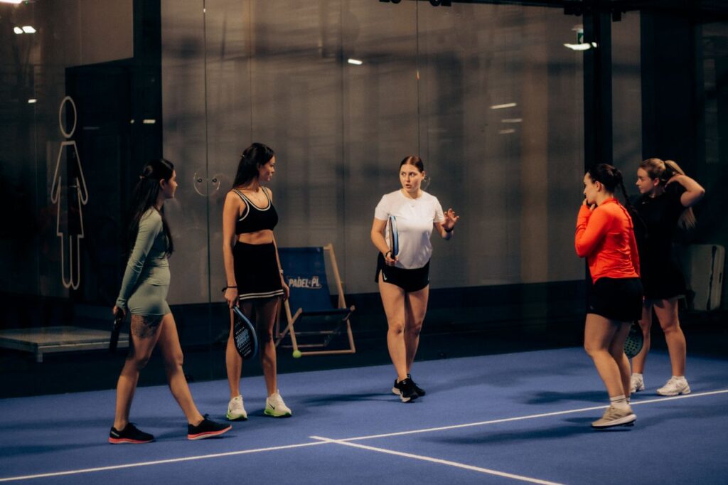 A group of female padel players chatting friendly on a blue indoor court, representing good padel etiquette and social play.