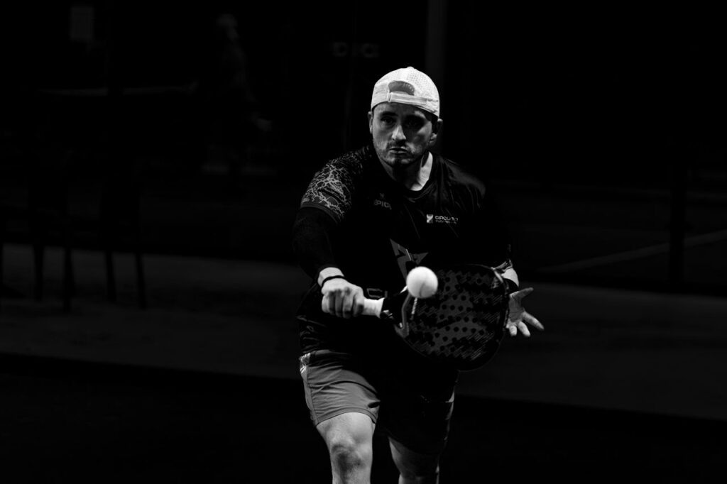 A dramatic black-and-white action shot of an intense male padel player striking the ball, highlighting the rigorous athleticism required by the IOC.