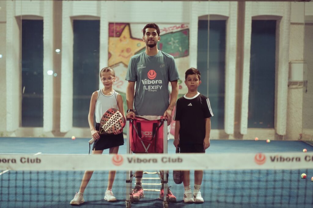 A padel coach standing at the net with two young students, representing a welcoming, inclusive community built on good court manners.