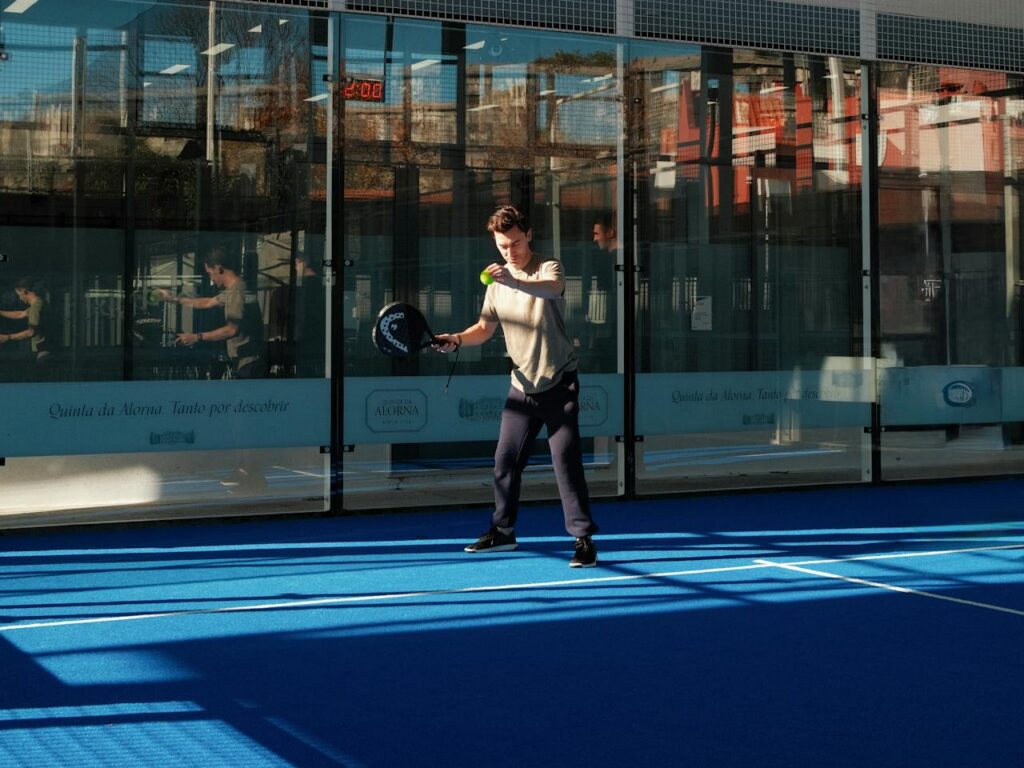 A male player in a beige t-shirt and dark pants tracks a ball near the glass walls of a bright, sunlit blue padel court.