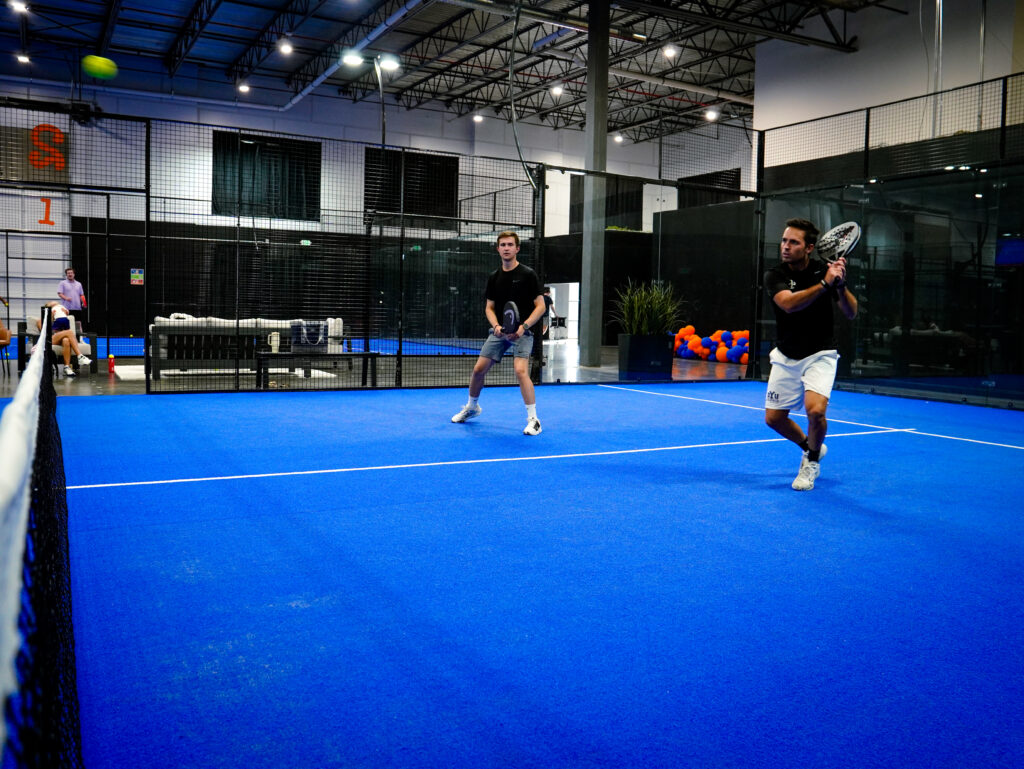 Two men playing an energetic game of indoor padel on a bright blue court, demonstrating a fun, active alternative to traditional gym workouts.