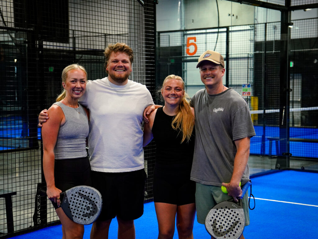 A group of four friends smiling and holding rackets together on an indoor padel court, highlighting the positive social benefits of group exercise.