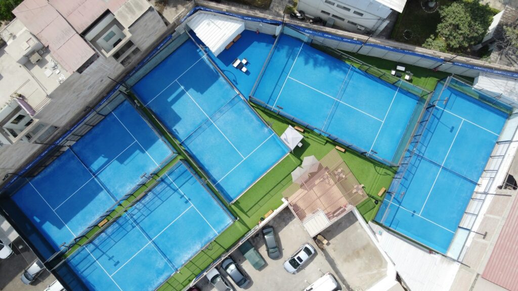 A high-angle view of an enclosed glass padel court surrounded by green trees, highlighting the court layout.