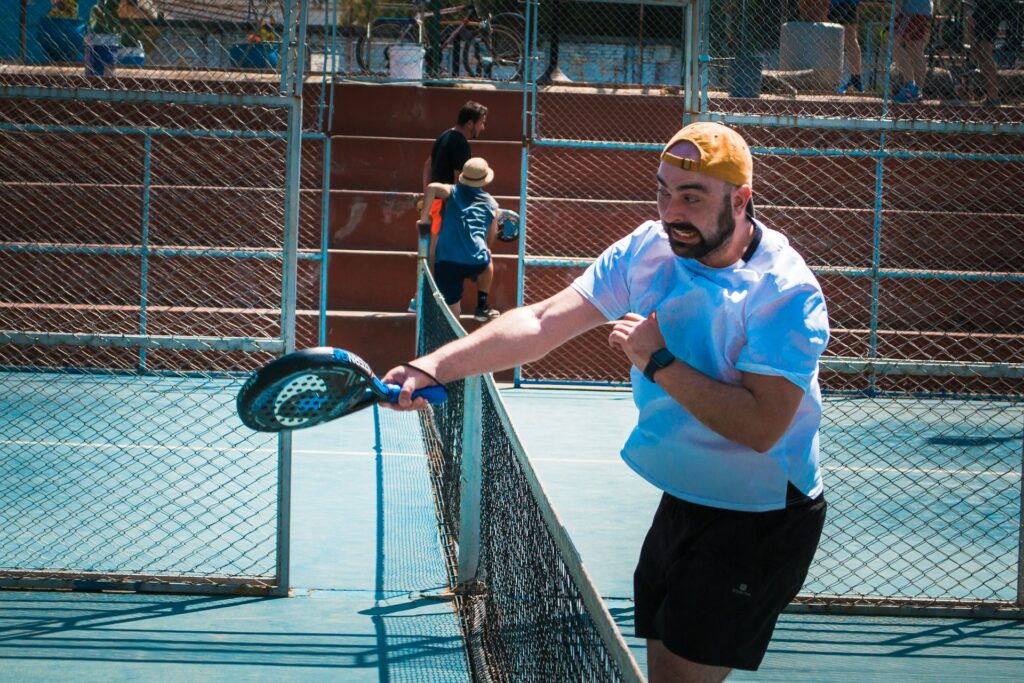 A man in a white t-shirt and yellow cap reaching his racket over the net during a match to demonstrate obscure padel rules.