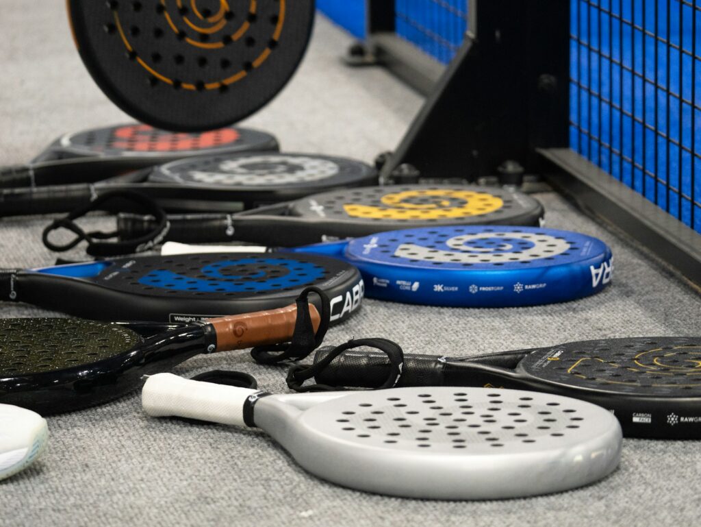 A collection of different padel rackets lying on the ground next to a court fence, showcasing the solid face and hole patterns that differentiate them from stringed squash rackets.
