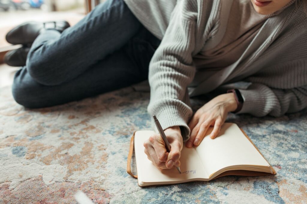 A woman lying comfortably on a rug writing in a blank notebook, representing the mental health benefits of starting a gratitude journal.