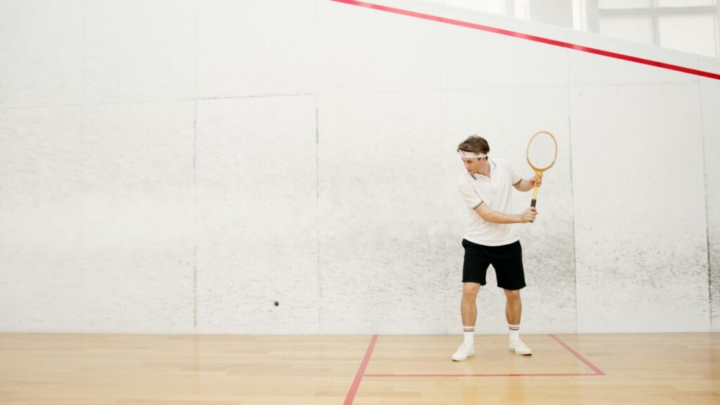A man in retro athletic wear holding a strung racket on a white indoor court, illustrating racquetball court setup and equipment.