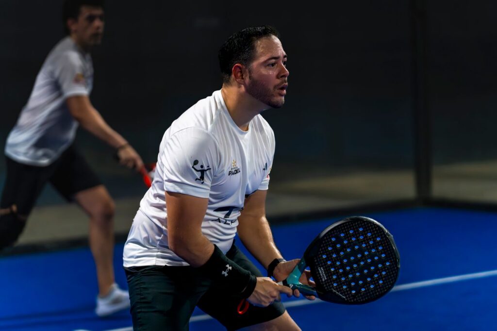 A focused player in a white shirt holding a solid perforated padel racket in a ready position on a blue indoor court.