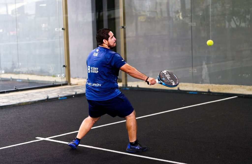 A focused male padel player in a blue shirt hitting an athletic attack shot on an outdoor court, demonstrating competitive doubles strategy.