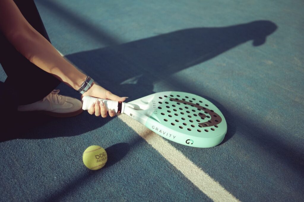 A close-up shot of a player picking up a mint green Head Gravity padel racket off a blue court surface next to a ball.