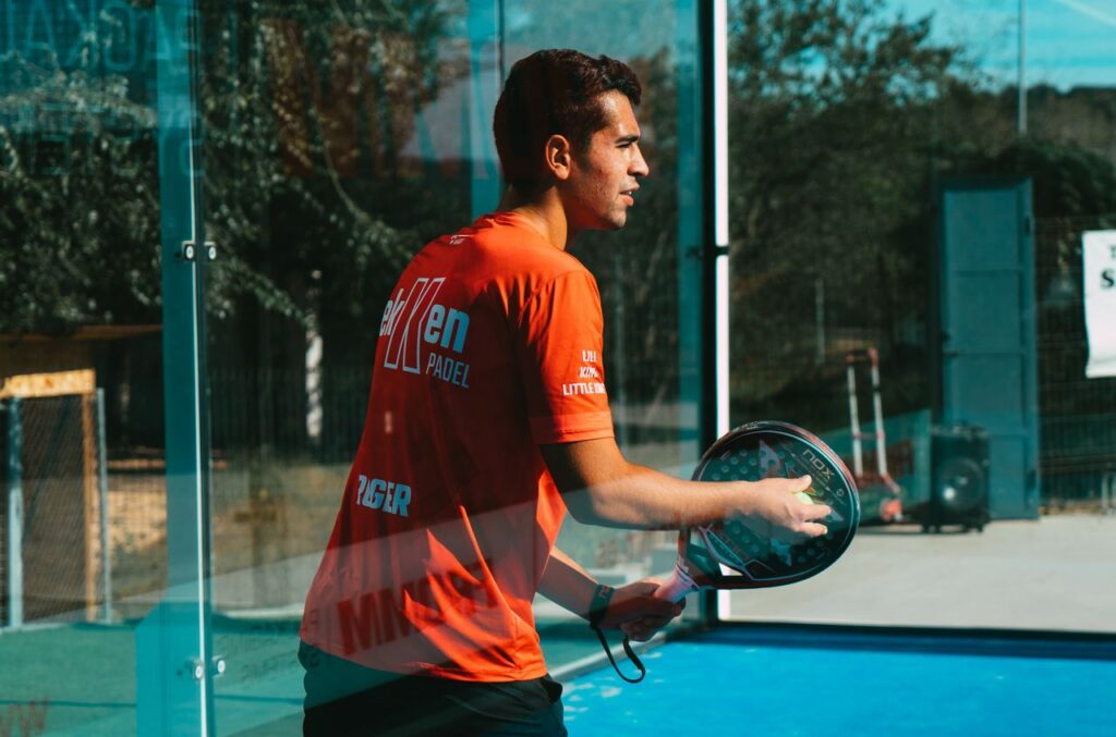 A padel player in an orange athletic shirt holding a racket near the glass wall, highlighting court boundary rules.