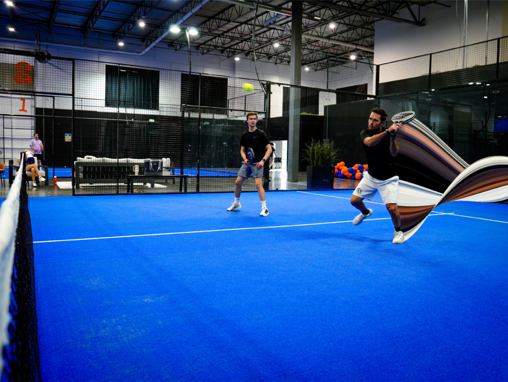 Two men playing a doubles padel match on a bright blue indoor turf court enclosed by glass walls and metal mesh fencing.