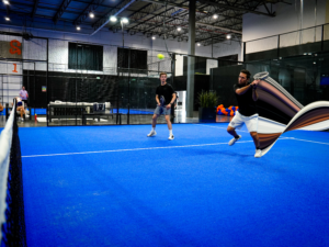 Two men playing a doubles padel match on a bright blue indoor turf court enclosed by glass walls and metal mesh fencing.