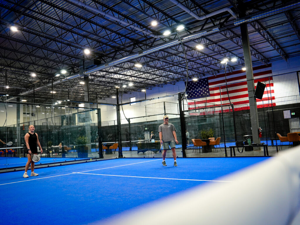 A man and a woman playing a game of padel on a bright blue indoor court. A large American flag hangs on the wall in the background of the facility.