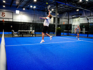 A male padel player jumping mid-air to hit an overhead smash on a blue indoor turf court while his female partner watches in the background.