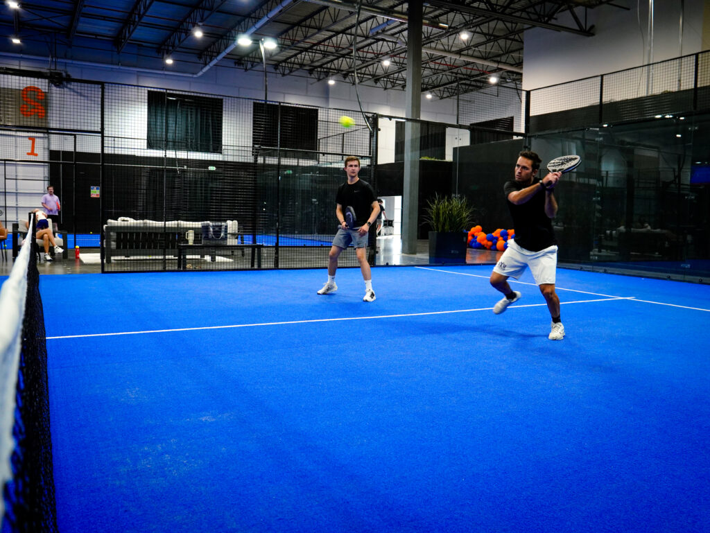 Two men playing an energetic doubles padel match on a bright blue indoor turf court at Padel Den in Utah.