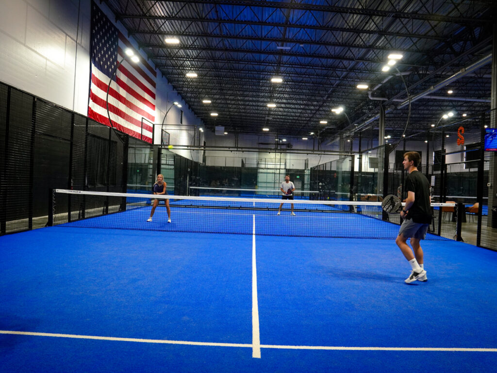 Four people playing a mixed doubles padel match on bright blue indoor turf courts beneath a large American flag at Padel Den in Orem, Utah.