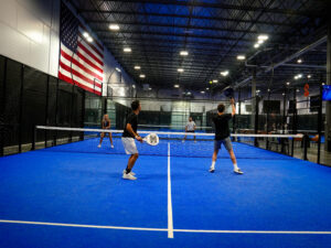 Four young adults playing an energetic doubles padel match on a bright blue indoor court beneath a large American flag during a group activity.