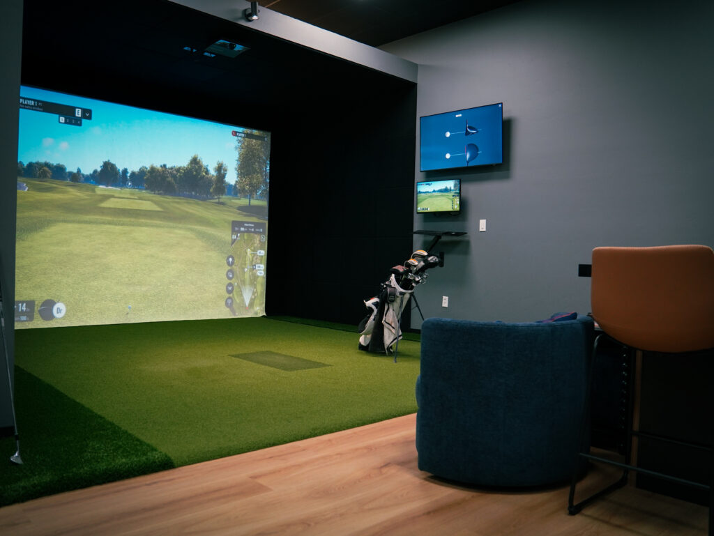 An indoor golf simulator bay with a large projection screen displaying a virtual fairway, golf clubs, and comfortable lounge seating.