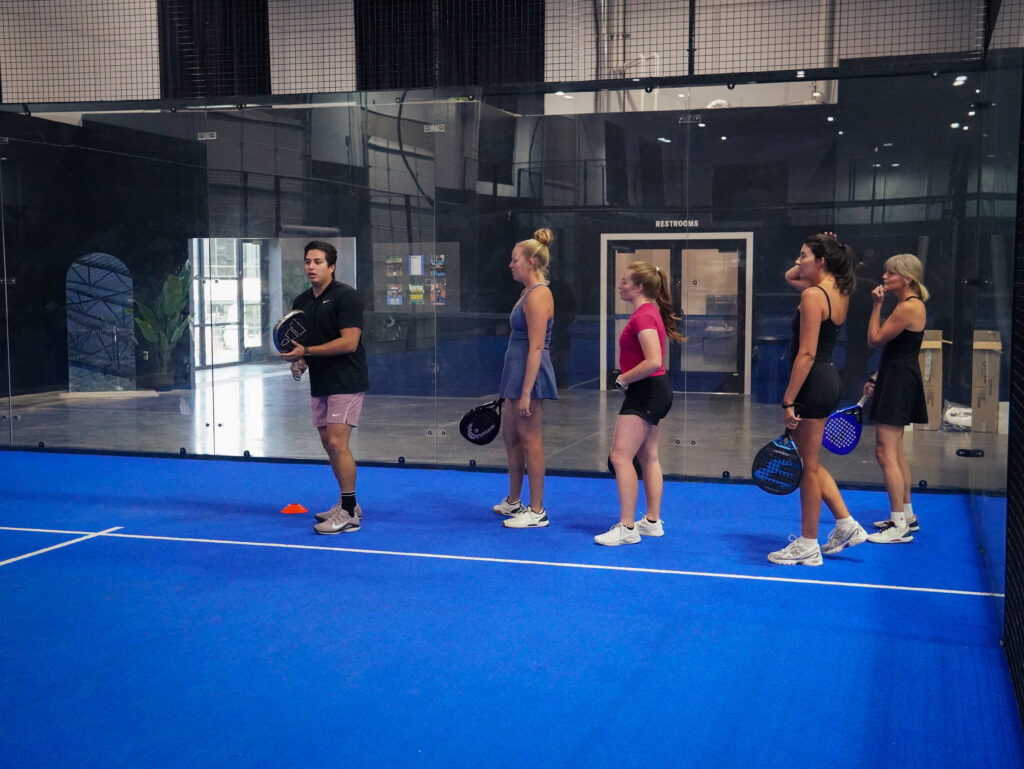 A male coach demonstrating technique to a group of four women holding rackets during a padel clinic on a blue indoor court at Padel Den.