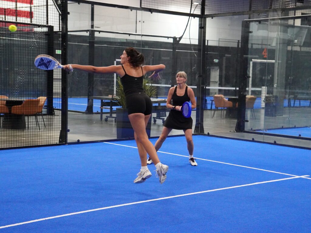 A woman leaping to hit a padel ball on a blue indoor court while her doubles partner watches, surrounded by glass walls and a lounge area.