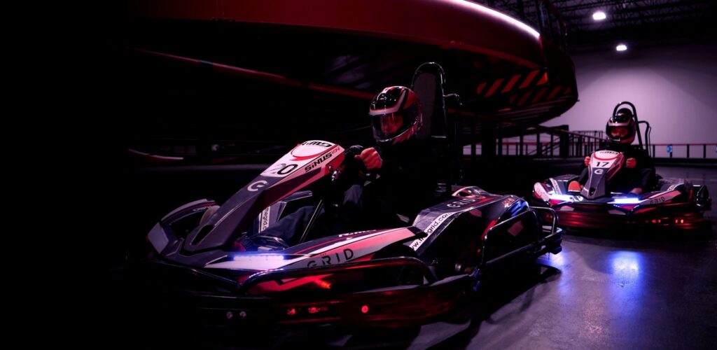 Drivers wearing helmets racing high-speed electric go-karts on an illuminated indoor track during an event.