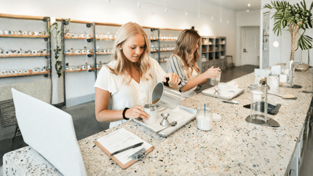 Two women standing at a fragrance bar pouring hot wax to create custom-scented candles during a group outing at The Wick Lab.